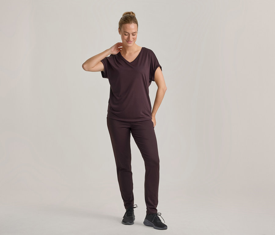 Frau in dunkelbraunem V-Ausschnitt-T-Shirt und passender Hose, mit schwarzen Turnschuhen, steht und blickt nach unten, eine Hand am Nacken.