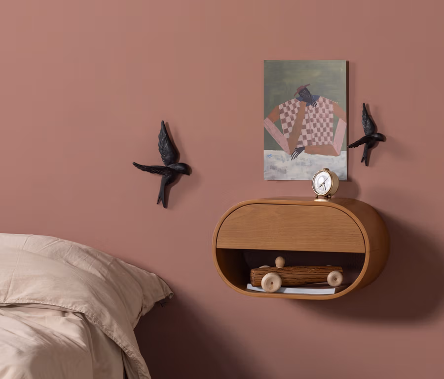 Eine staubrosa Schlafzimmerwand mit einem hellen, ovalen Holz-Wandregal. Auf dem Regal stehen ein goldener Wecker und ein Gemälde einer Figur. Zwei schwarze Vogeldekorationen flankieren das Regal. Ein hölzernes Spielzeugauto liegt im offenen Fach des Regals. Links ist ein Bett mit beiger Bettwäsche zu sehen; auf dem Boden ein dunkelbrauner, kugelförmiger Pouf.