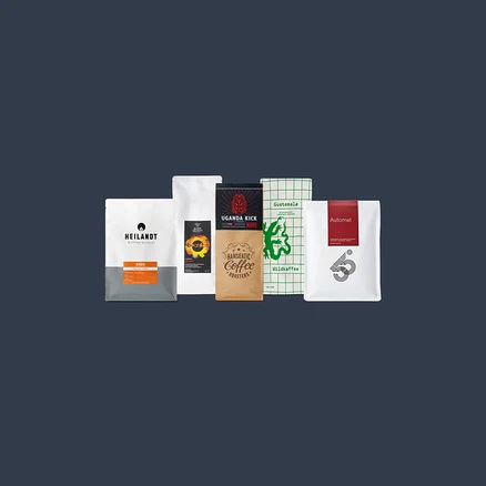 Fünf aufrecht stehende Kaffeepackungen: eine weiß-graue „HEILANDT ENZO“-Packung, eine weiße „BLACK DELIGHT beanice“-Packung, eine braune „UGANDA KICK HANSEATIC COFFEE ROASTERS“-Packung, eine weiße „Guatemala Wildkaffee“-Packung mit grünem Krokodil und eine weiße „Automat 65°“-Packung mit rotem Streifen.