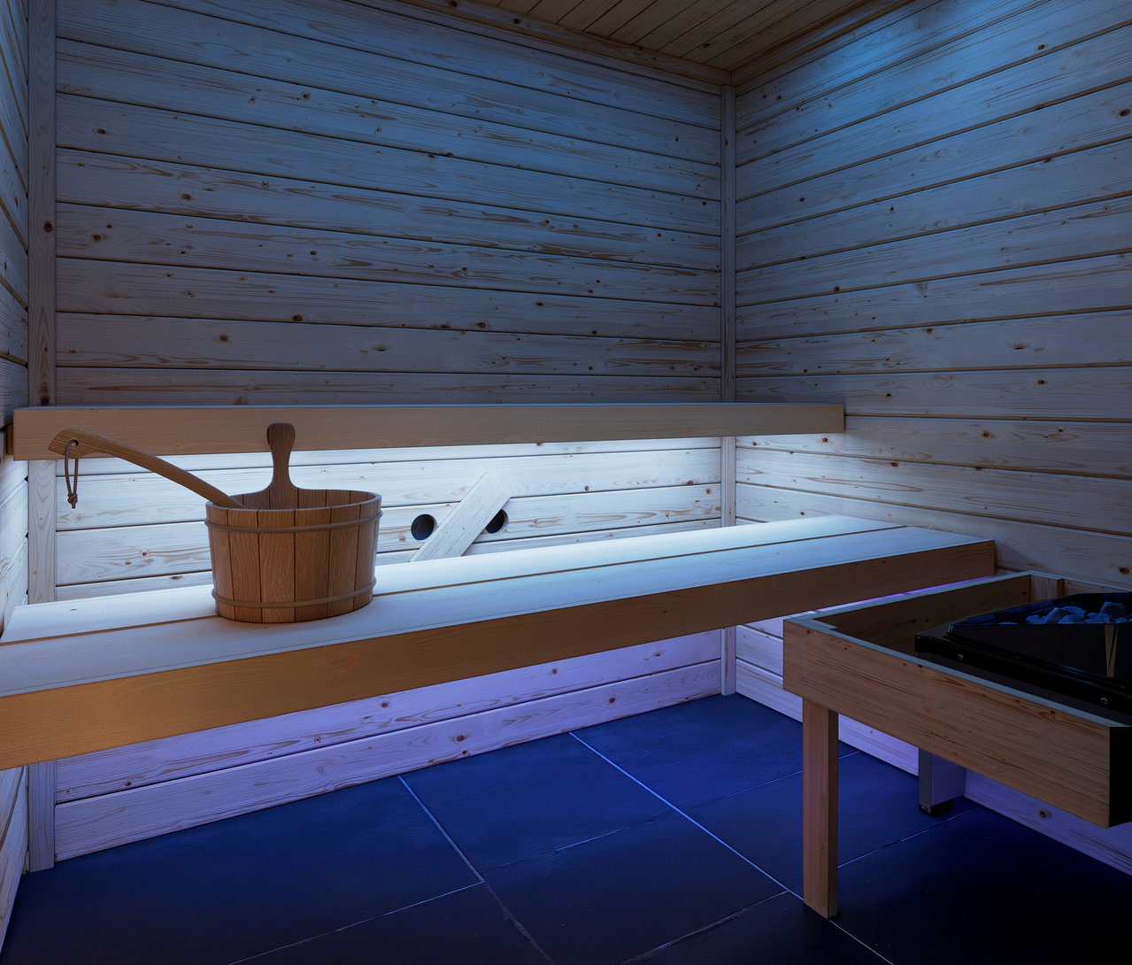 Sauna-Innenraum mit hellen Holzwänden und -bänken, einem Holzeimer und einer Schöpfkelle auf einer Bank. Blaue LED-Beleuchtung unter den Bänken, rechts ein Saunaofen mit Steinen auf dunklem Fliesenboden.