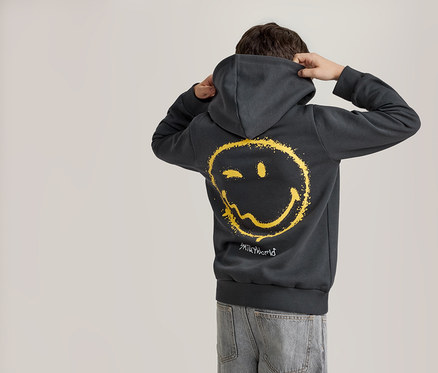 Eine Person von hinten, die einen dunkelgrauen Hoodie mit einem verwaschenen gelben Smiley-Gesicht auf dem Rücken und hellblaue Jeans trägt. Die Person hält die Kapuze mit beiden Händen.