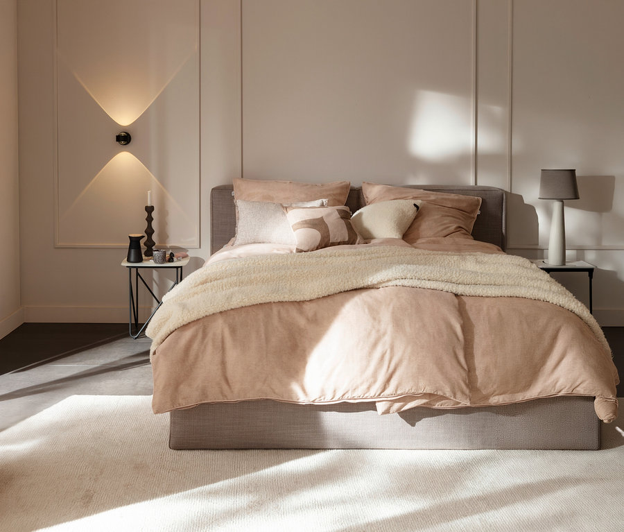 Ein Schlafzimmer mit einem Bett, das mit rosa Bettwäsche und einer offwhite SCHÖNER WOHNEN-Kollektion Wohndecke »TEDDY« bezogen ist. Links vom Bett steht ein Nachttisch mit Kerze und Vase unter einer Wandleuchte. Rechts steht ein Nachttisch mit einer Tischlampe. Sonnenlicht fällt auf den hellen Teppich vor dem Bett.