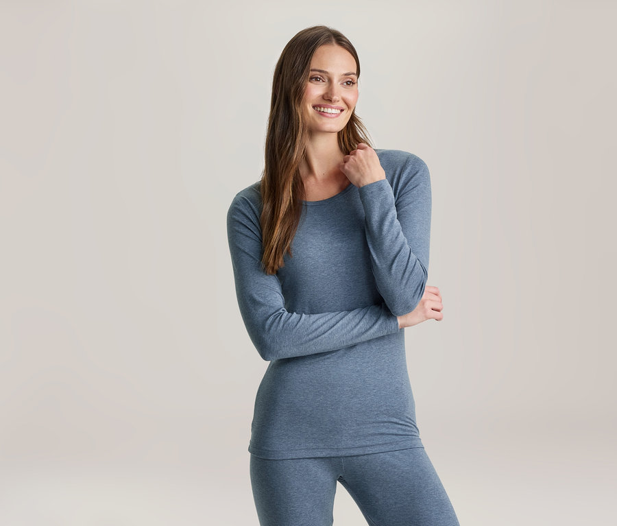 Eine lächelnde Frau mit langen braunen Haaren trägt ein blaues Langarmshirt und passende Leggings und blickt nach rechts.