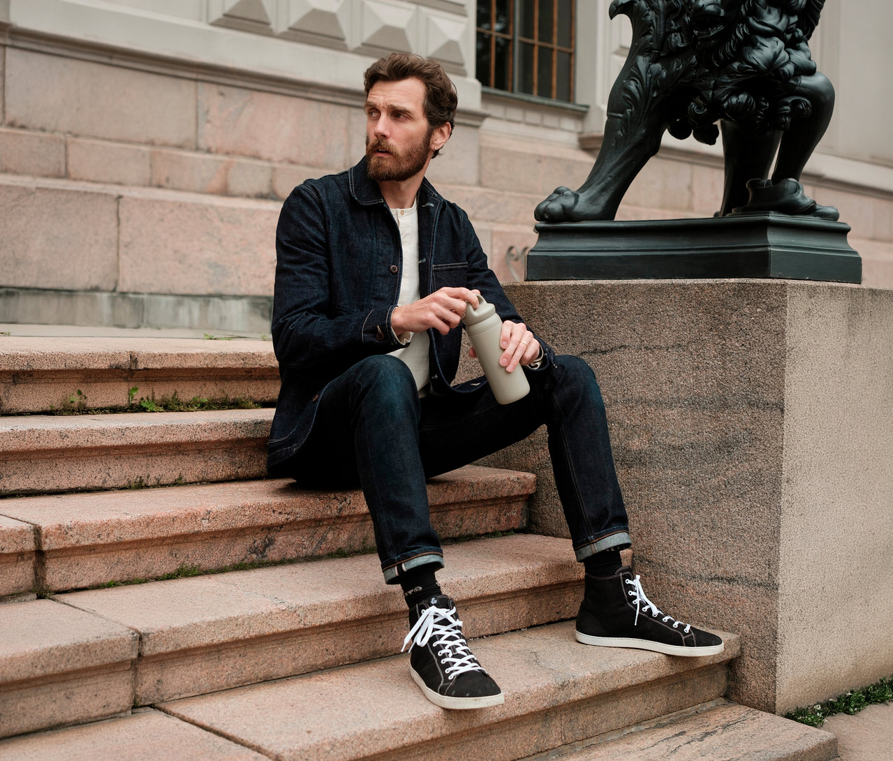 Mann mit Bart sitzt auf Steinstufen im Freien und hält einen beigen KINTO Tumbler »Day Off«. Er trägt eine Jeansjacke und Jeans.