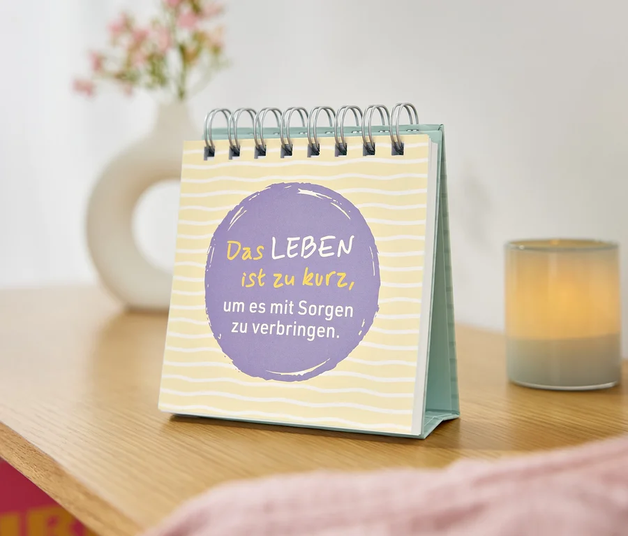 Tischkalender mit dem Zitat "Das Leben ist zu kurz, um es mit Sorgen zu verbringen" vor einer Vase und einer Kerze.
