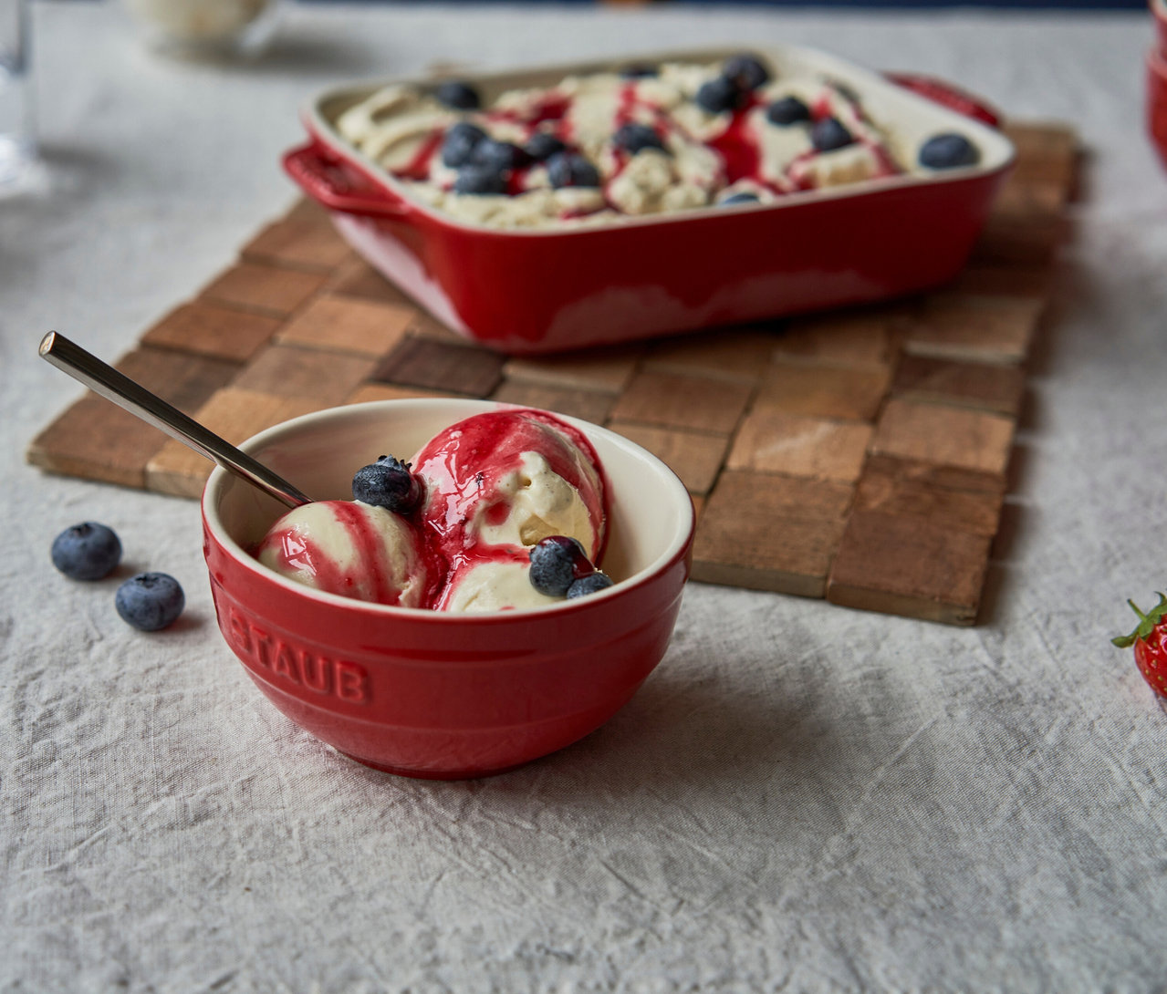 Kirschrote STAUB Keramik-Schüssel mit Vanilleeis, roter Soße und Blaubeeren; im Hintergrund eine Auflaufform mit Dessert.