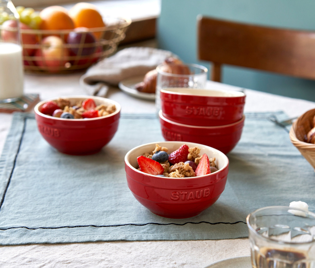 Frühstückstisch mit zwei kirschroten STAUB Keramik-Schüsseln, gefüllt mit Müsli, Erdbeeren und Heidelbeeren. Daneben sind zwei weitere kirschrote STAUB Keramik-Schüsseln gestapelt. Im Hintergrund ein Obstkorb mit Orangen, Trauben und Äpfeln, daneben ein Glas Milch und Croissants.