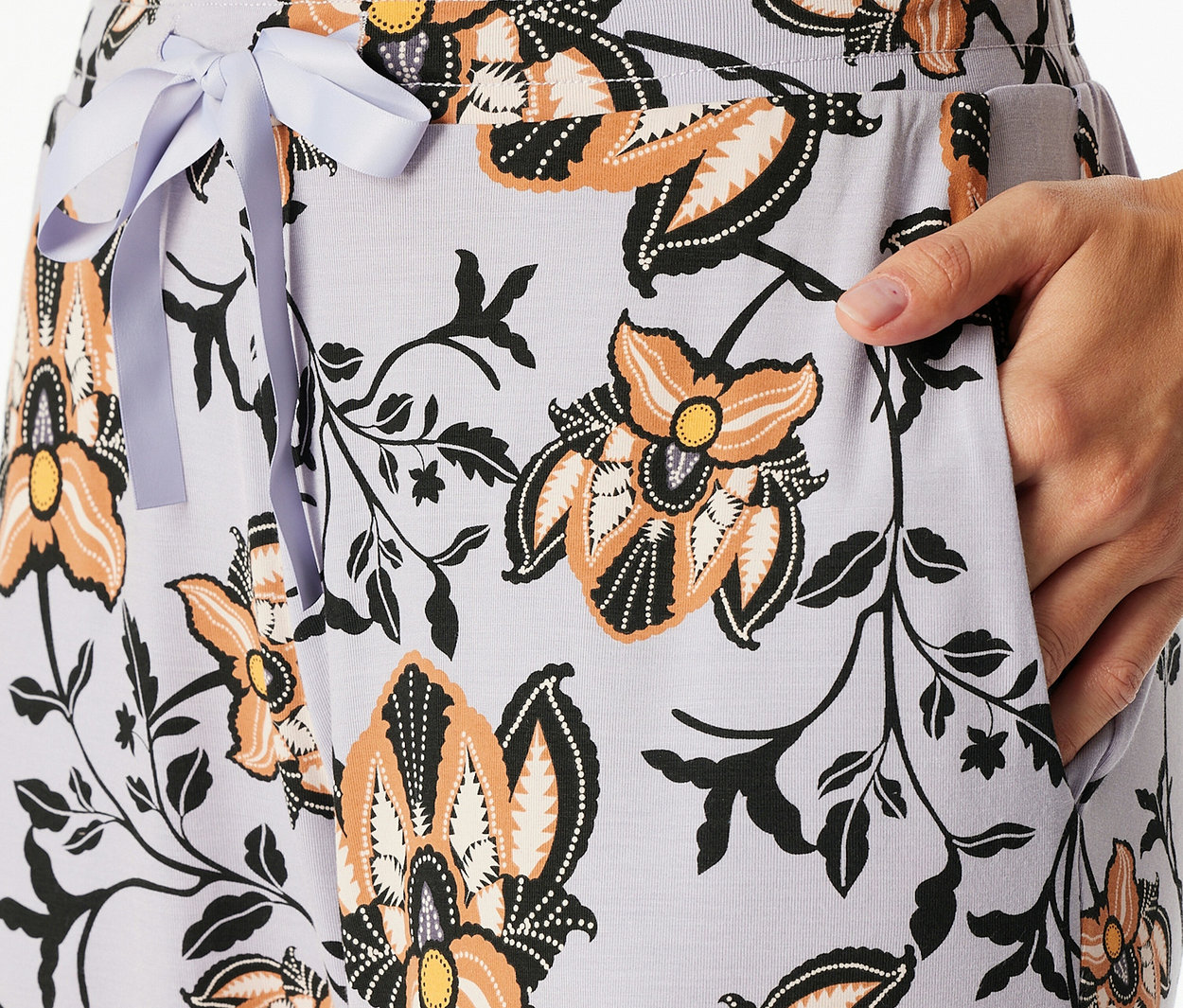 Nahaufnahme einer hellvioletten Hose mit orangefarbenem, schwarzem und weißem Blumenmuster, mit einer hellvioletten Schleife an der Taille und einer Hand in der Tasche.