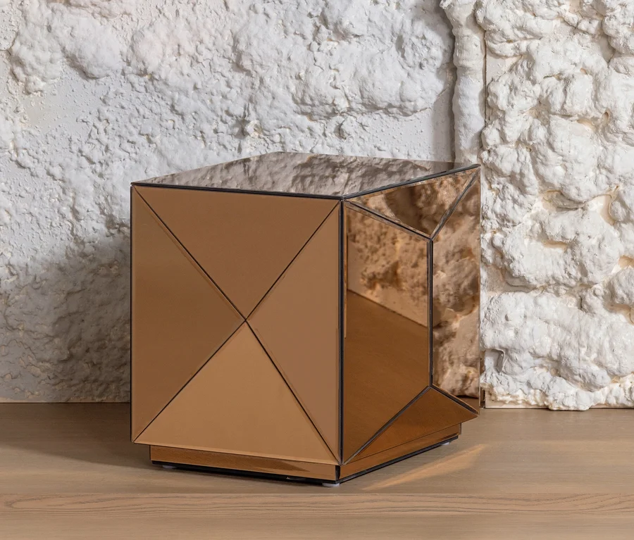 Ein quadratischer, roségoldener Spiegeltisch mit geometrischem, facettiertem Design steht auf einem hellen Holzfußboden vor einer strukturierten weißen Wand.
