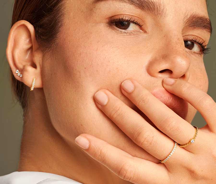 Porträt einer Frau, die ihre Hand ans Gesicht hält. Sie trägt einen 585 Gold Ring Eternity Zirkonia und einen 585 Gold Ring Kugeln und Zirkonia. Sie trägt 585 Gold Ohrstecker Zirkonia Verlauf.