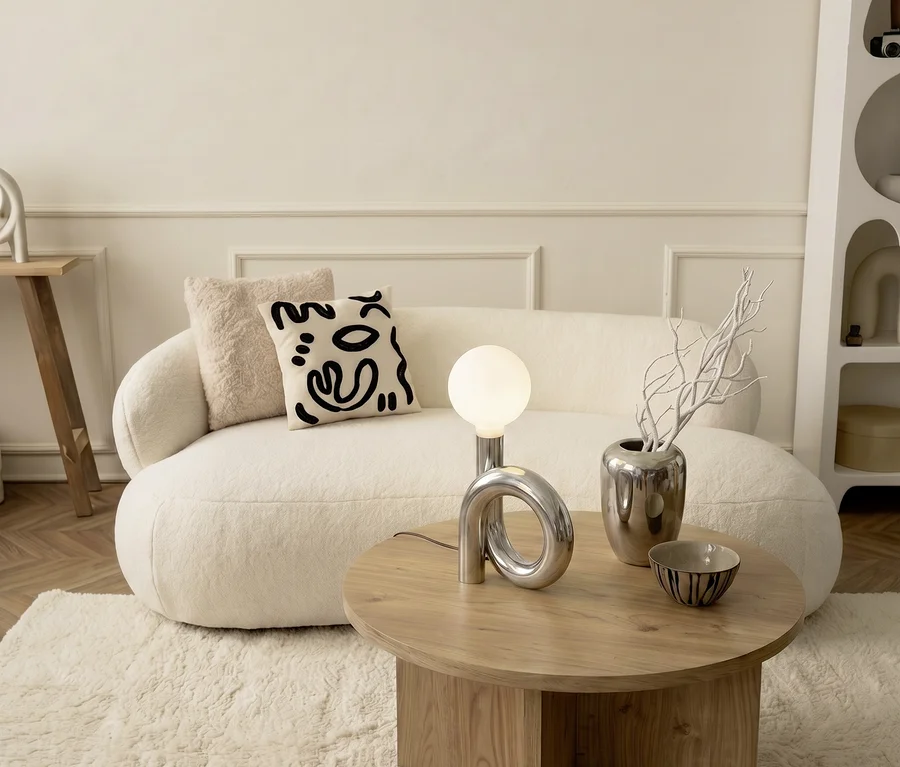 Ein cremefarbenes Sofa mit zwei Kissen, eines schlicht, eines mit abstraktem schwarzem Gesichtsmuster, steht auf einem flauschigen cremefarbenen Teppich. Vor dem Sofa steht ein runder Holztisch mit einer modernen silbernen Lampe, einer silbernen Vase mit weißen Ästen und einer kleinen gemusterten Schale.