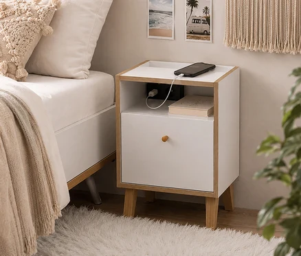Weißer Nachttisch mit Holzrahmen neben einem Bett. Ein Smartphone lädt auf der Ablage, darunter ein Buch und ein Ladegerät. Das Bett hat weiße Bettwäsche, eine beige Decke und ein Kissen. Ein flauschiger Teppich liegt auf dem Boden.