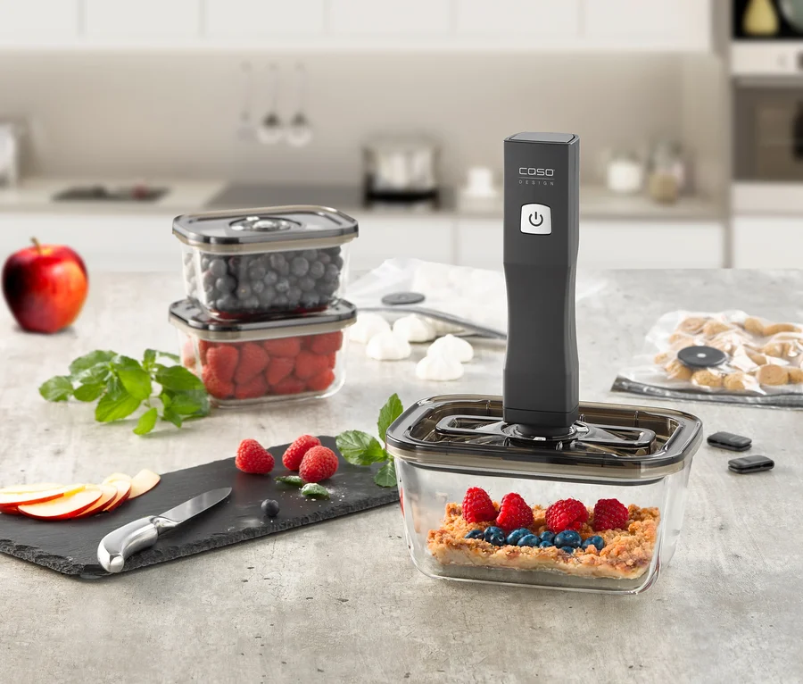 Ein CASO Vacu OneTouch Handvakuumierer vakuumiert einen Behälter mit Beeren-Crumble auf einer Küchentheke, daneben stehen zwei gestapelte Behälter mit Heidelbeeren und Himbeeren, Apfelscheiben und Minze liegen auf einer Schieferplatte.
