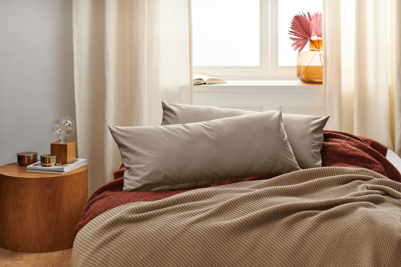 Zwei beige Perkal-Kopfkissenbezüge liegen auf einem Bett mit einer braunen Decke.