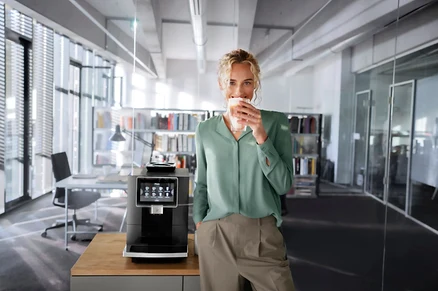 Blonde Frau im grünen Oberteil trinkt Kaffee neben einer Kaffeemaschine im Büro.