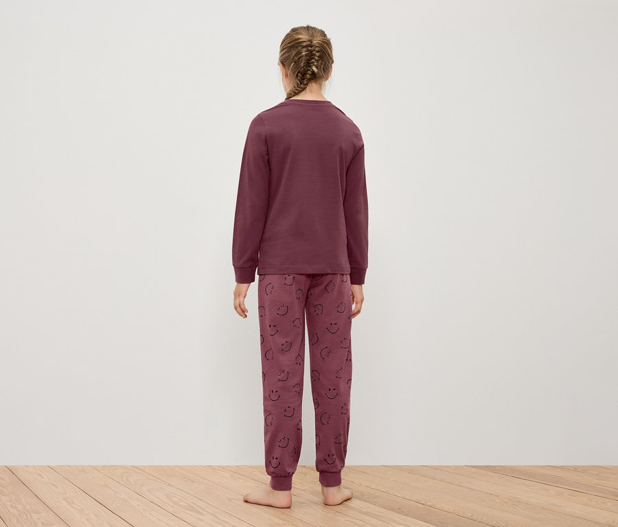 Mädchen in bordeauxfarbenem Langarm-Schlafanzug und Hose mit Smiley-Gesichtern, steht mit dem Rücken zum Betrachter auf einem Holzboden vor einer weißen Wand.