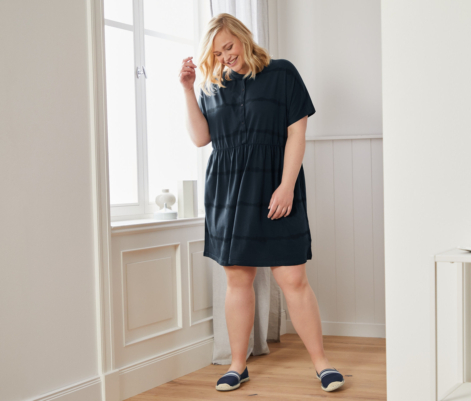 Frau steht am Fenster und trägt ein Jerseykleid und Espadrilles.