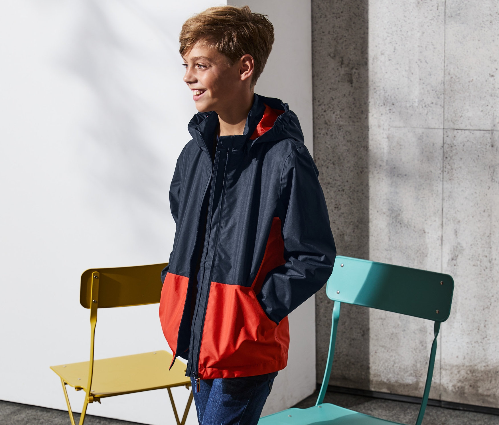 Ein Junge trägt eine Regenjacke und steht neben einem gelben und einem türkisfarbenen Stuhl.