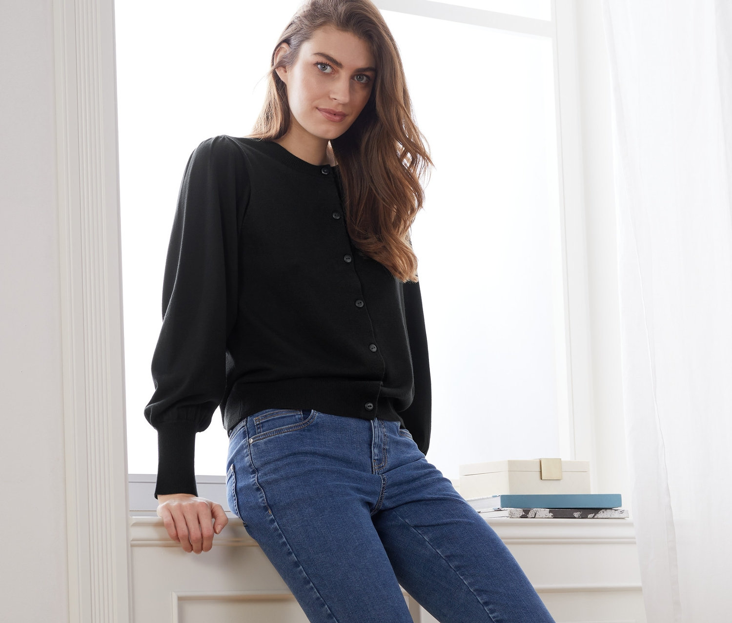 Frau sitzt auf Fensterbank, trägt Feinstrickjacke mit Puffärmeln und blaue Jeans.