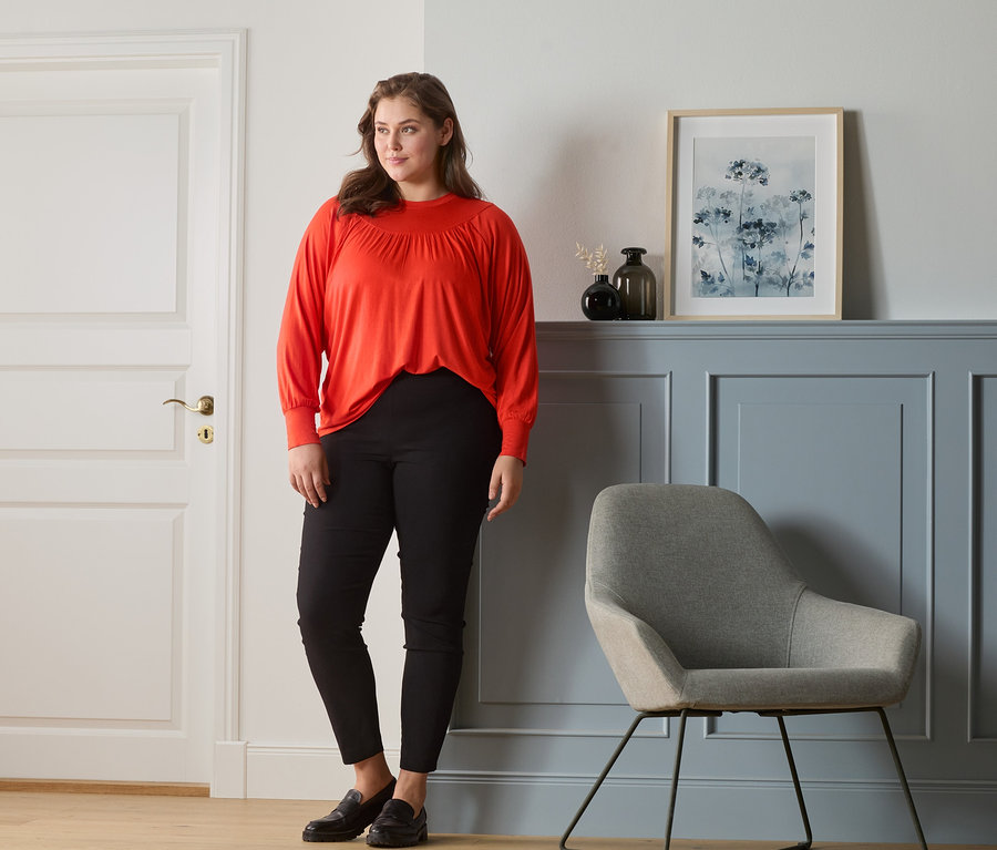 Frau steht und trägt ein orange-rotes Blusenshirt mit Raffung und eine Stretchhose.