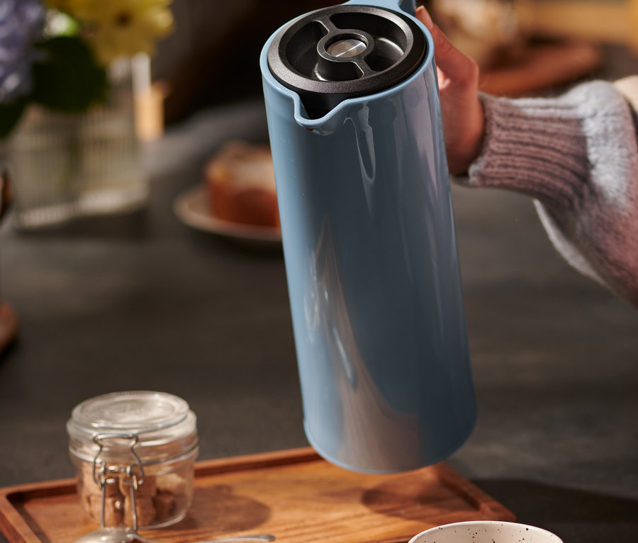 Hand hält blaue Isolierkanne über Tasse und Holztablett.