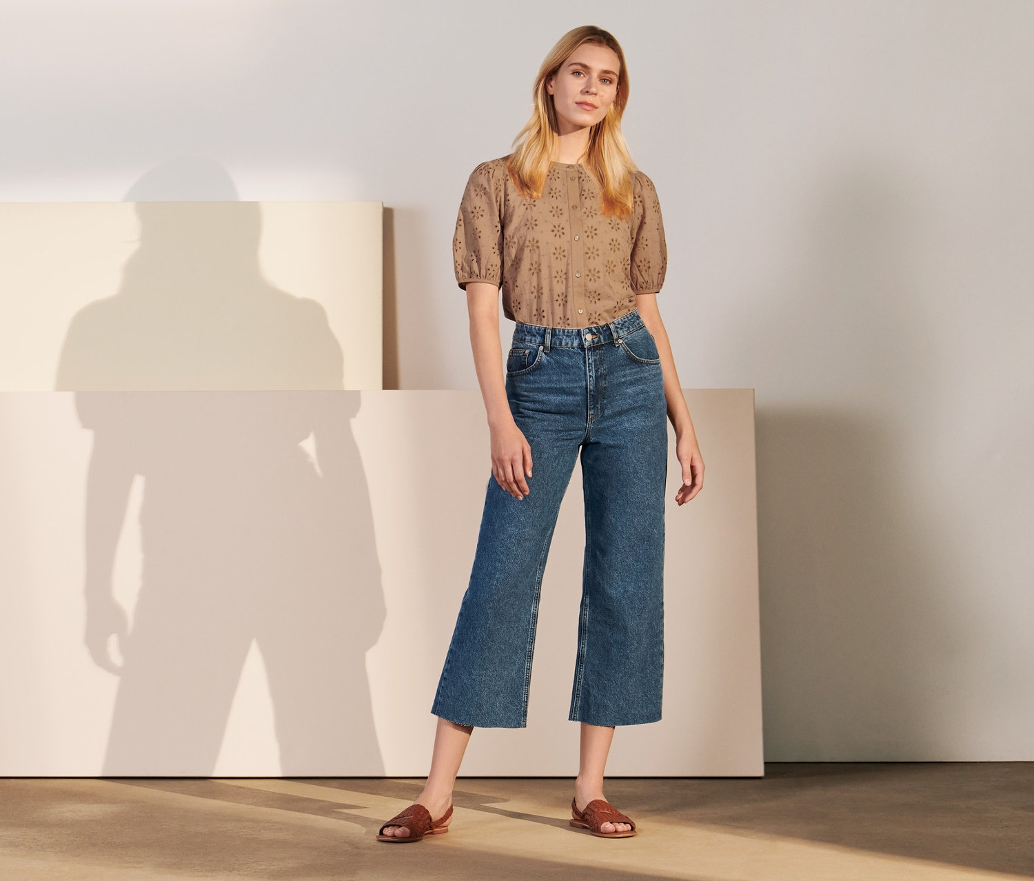 Eine Frau posiert stehend in Culotte-Jeans und brauner Bluse.