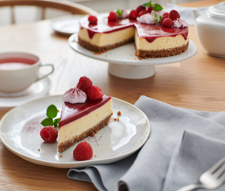 Auf einem Tisch stehen ein Teller mit einem Stück Kuchen, das mit Himbeeren und Minze verziert ist. Im Hintergrund befindet sich der Rest des Kuchens auf einem Tablett, eine Tasse Tee und eine Teekanne. Tortenheber