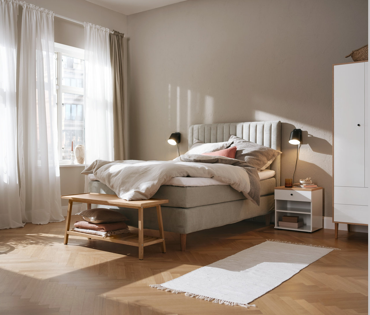 Beiges Boxspringbett (ca. 180 x 200 cm), Kleiderschrank, Nachttisch namens »Bekeson«, schwarze Wandleuchte aus Metall und Sitzbank mit Ablage.