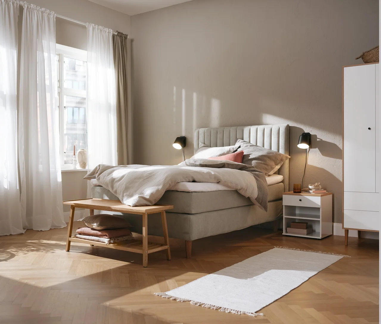 Beiges Boxspringbett (ca. 180 x 200 cm), Kleiderschrank, Nachttisch namens »Bekeson«, schwarze Wandleuchte aus Metall und Sitzbank mit Ablage.