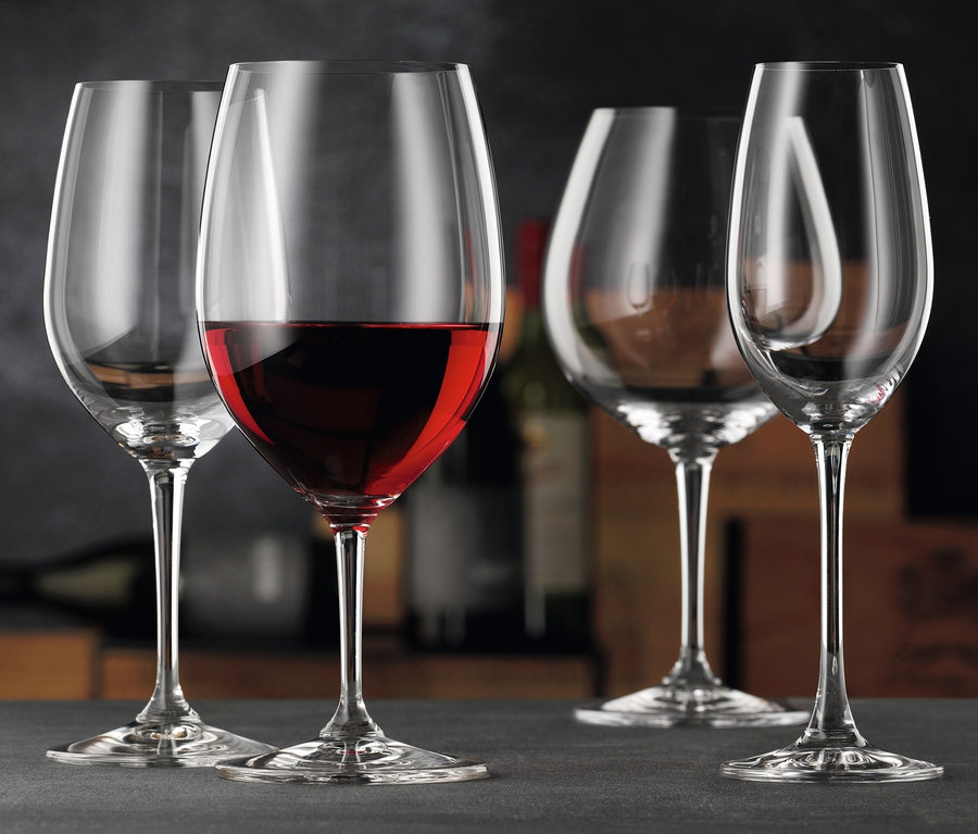 Vier Weingläser, eines mit Rotwein gefüllt, stehen auf einer grauen Oberfläche. Im Hintergrund sind weitere Flaschen und Kisten zu sehen. Dabei sind 8 Weißweingläser »Nachtmann ViVino«, 8 Champagnergläser »Nachtmann ViVino« und 8 Rotweingläser »Nachtmann ViVino«.