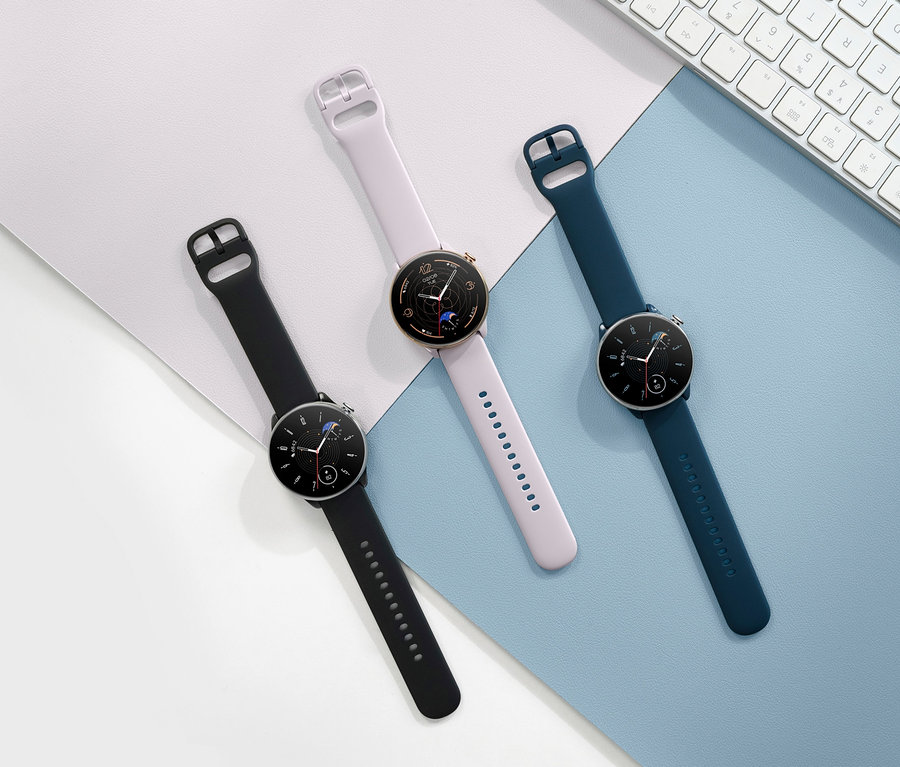 Drei Amazfit GTR Mini Smartwatches in Schwarz, Rosa und Blau liegen auf einer blau-weißen Oberfläche neben einer weißen Tastatur.