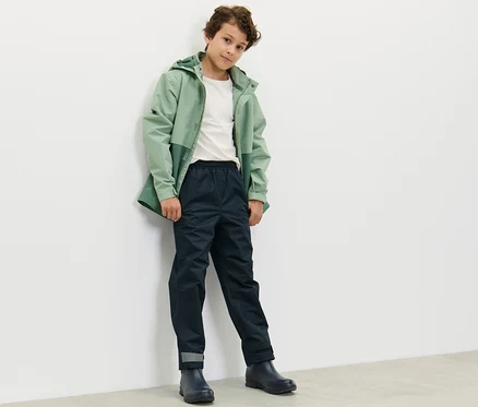 Ein Junge in einer grünen Jacke und dunkelblauer Kinder-Regenhose steht an einer weißen Wand.