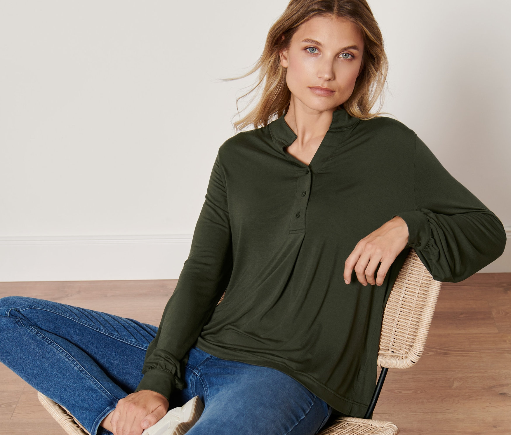 Eine Frau sitzt auf einem Stuhl und trägt ein Blusenshirt, Jeans und Turnschuhe.