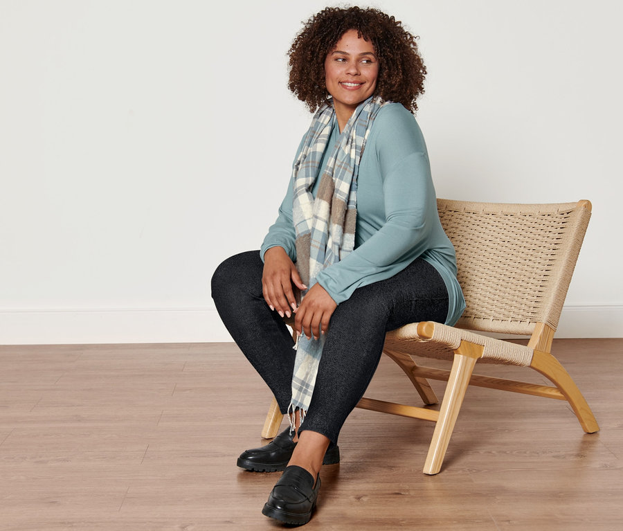 Eine Frau sitzt auf einem Stuhl und trägt einen karierten Webschal, eine gefütterte Leggings in Jeansoptik, ein rauchblaues Langarmshirt mit V-Ausschnitt und Leder-Loafer.