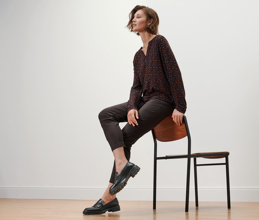 Frau sitzt auf einem Stuhl und trägt Leder-Loafer, eine Bengalin-Hose und ein bedrucktes Blusenshirt.