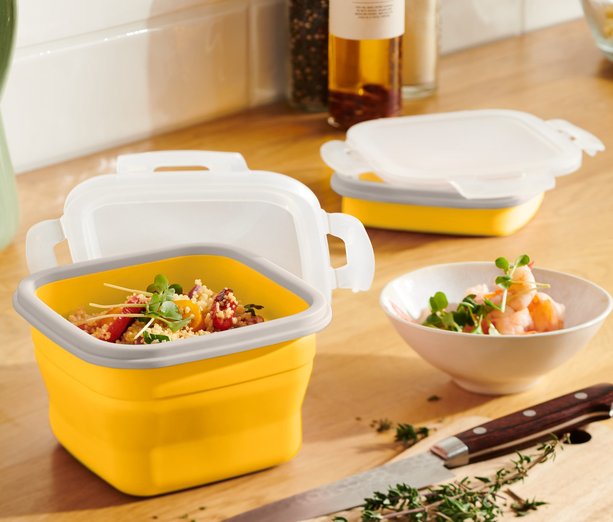 Eine gelbe, faltbare Frischhaltedose mit Inhalt steht auf einem Holztisch neben einem grünen Krug und einer Schale mit Garnelen.