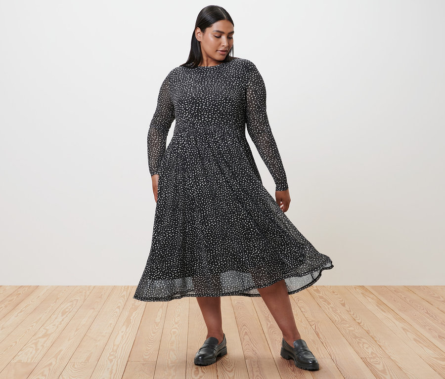 Frau in gepunktetem Mesh-Kleid und Leder-Loafern steht auf Holzboden.