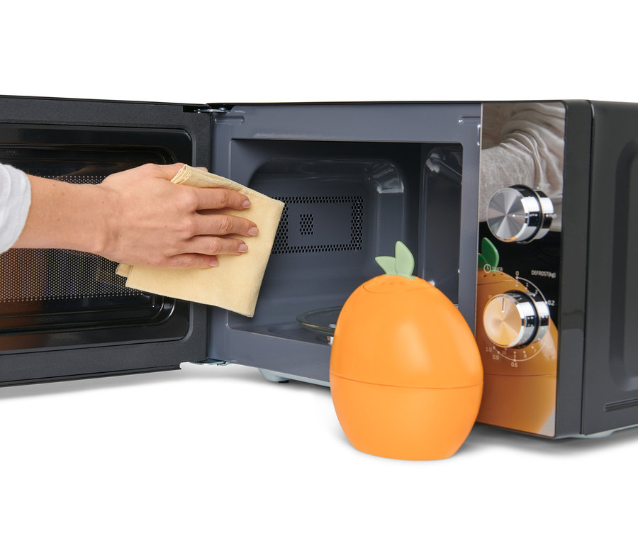 Hand wischt das Innere einer Mikrowelle mit einem Tuch neben einem Mikrowellenreiniger in Orangenform.