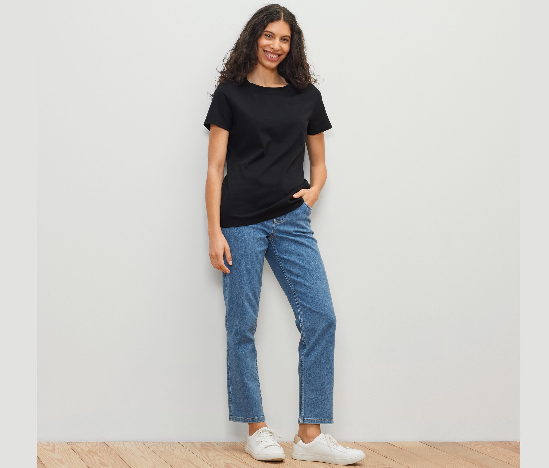 Frau mit schwarzem Basic T-Shirt, blauen Jeans und weißen Schuhen steht vor einer weißen Wand.