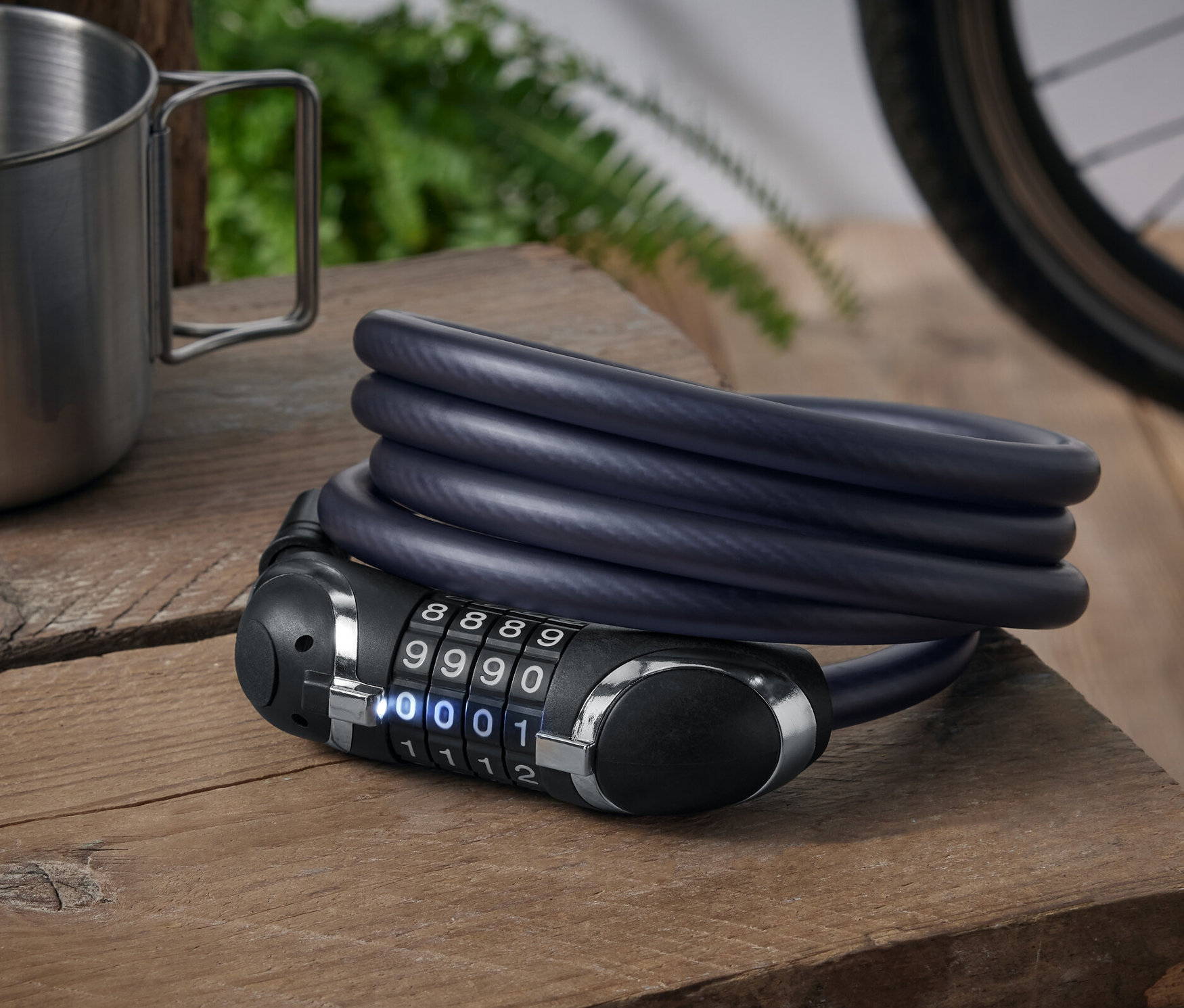 Auf einem Holztisch liegen ein Stahlbecher und ein aufgerolltes, extralanges Universal-Stahlkabelschloss mit Zahlencode.