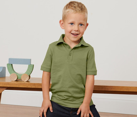 Ein Junge posiert auf einer Bank in einem olivfarbenen Poloshirt und Jeans. Auf der Bank liegen 2 Kleinkinder-Poloshirts.