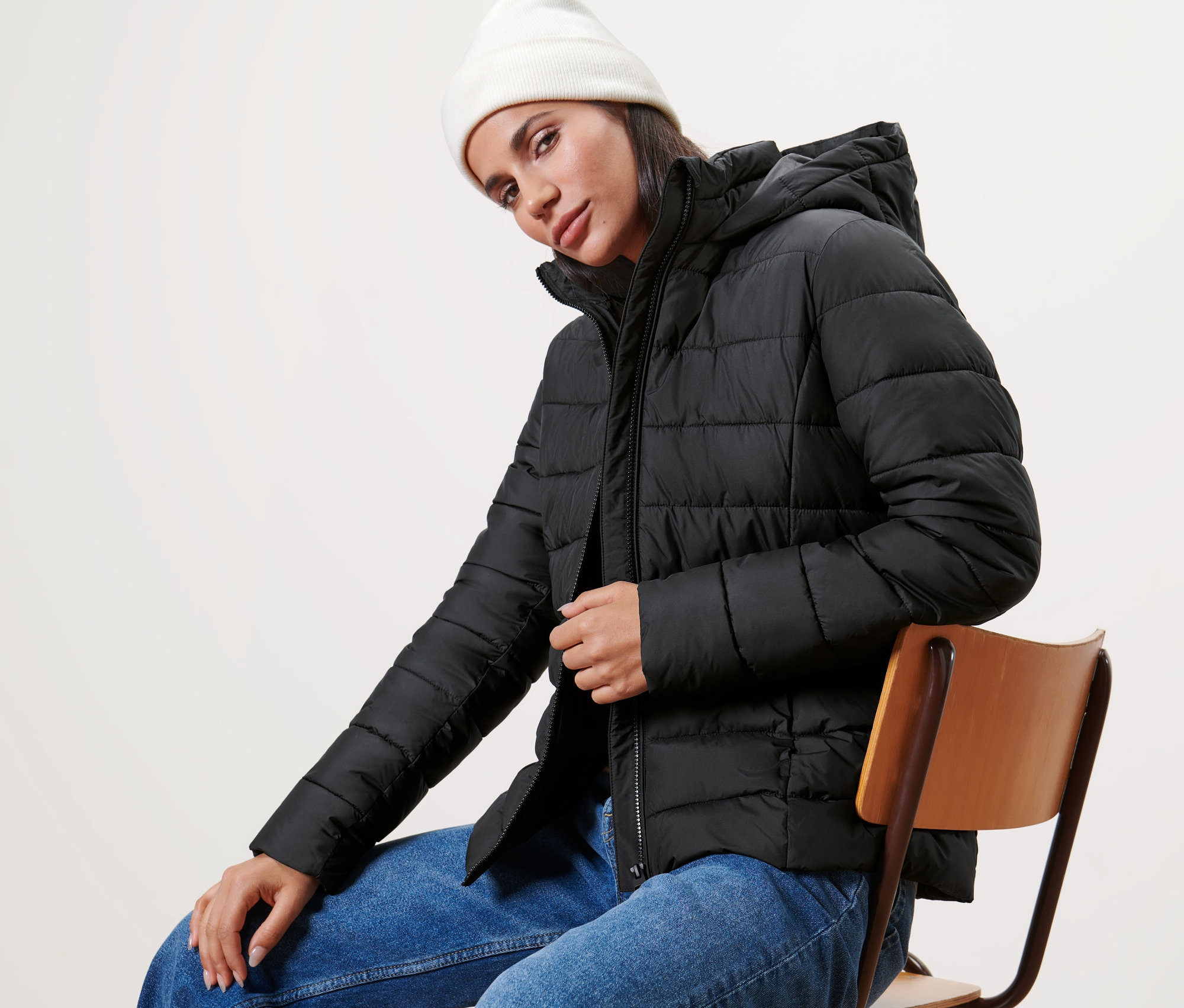 Frau sitzt auf einem Stuhl und trägt eine schwarze Steppjacke mit Kapuze und eine weiße Mütze.