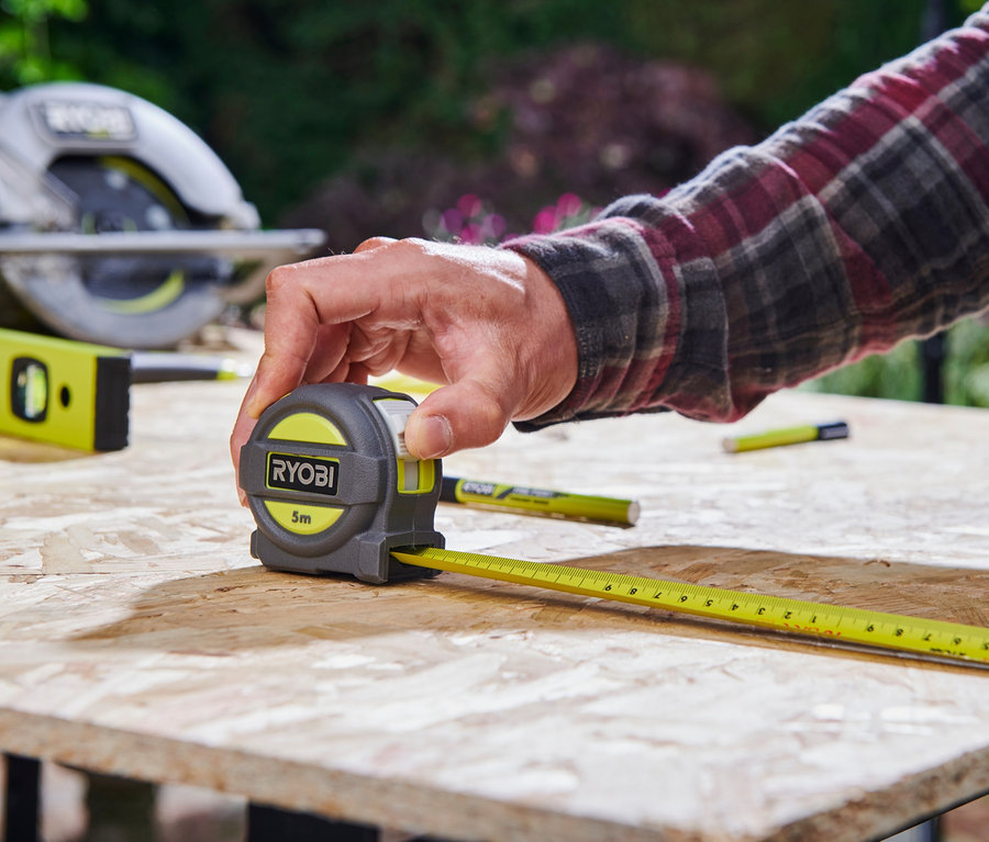 Mann misst ein Holzbrett mit einem Rollbandmaß. Im Hintergrund befindet sich ein Werkzeugset RYOBI 18 V ONE+.