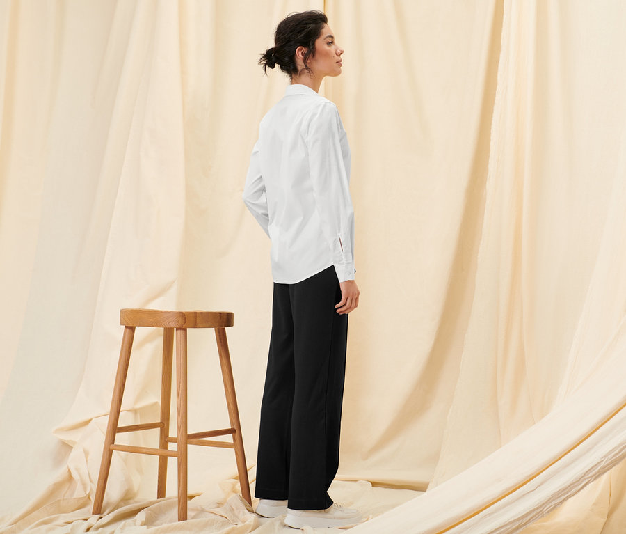 Eine Frau steht seitlich und trägt eine Hemdbluse und eine schwarze Sweathose.