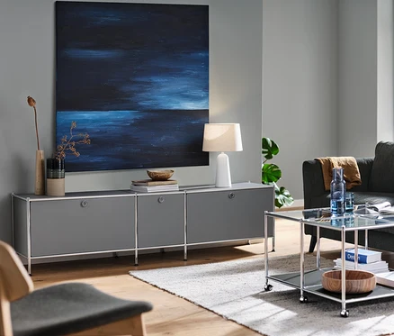 Graues Lowboard »CN3« mit 3 Klappen, eine Tischleuchte mit weißem Textilschirm und ein anthrazitfarbenes 3-Sitzer-Sofa in einem Wohnzimmer. Ebenfalls ein Couchtisch mit Rollen »CN3« und ein Loungechair.