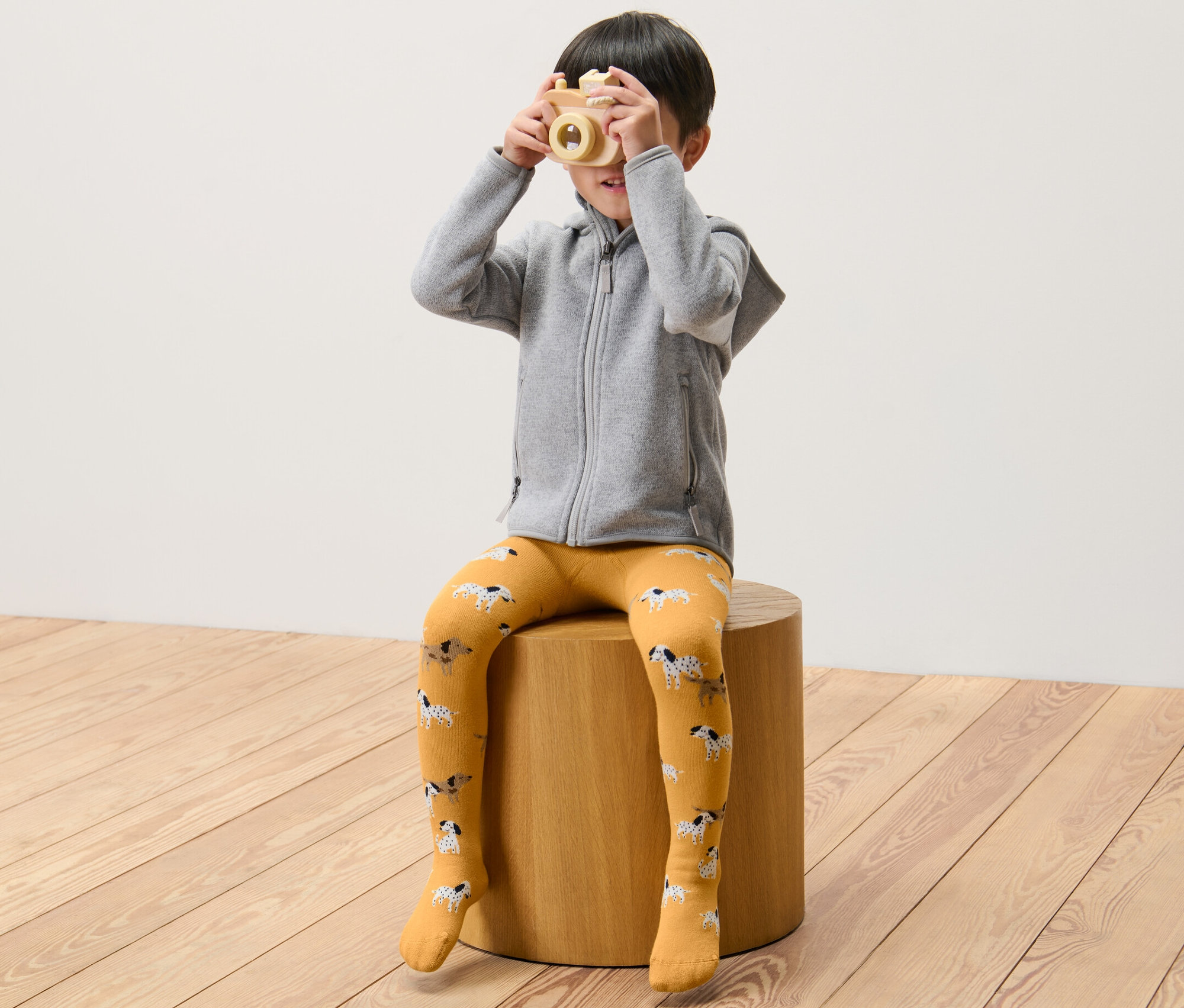 Ein Junge sitzt auf einem Hocker und hält eine Holzkamera vor sein Gesicht. Er trägt eine grüne und gelbe Kinder-Thermostrumpfhose.
