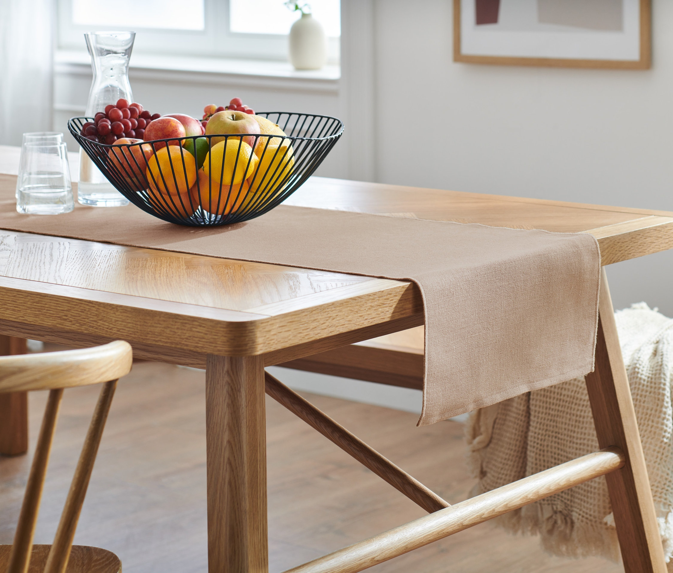 Auf einem Holztisch liegt ein beiger Tischläufer. Darauf steht eine Drahtschale mit Obst.