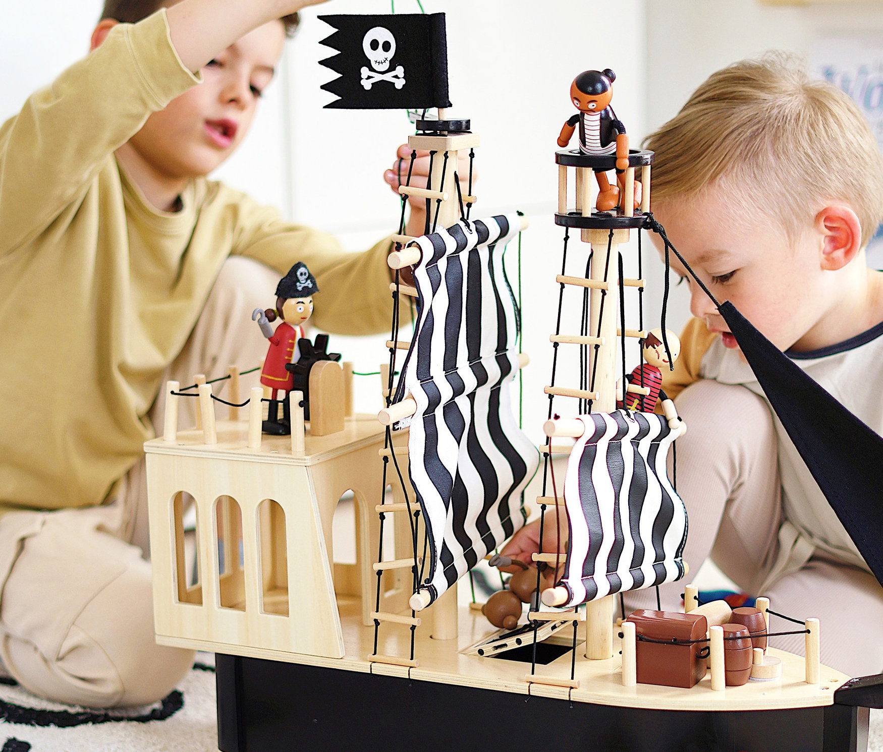 Zwei Jungen spielen mit einem „small foot Piratenschiff“ aus Holz auf einem Teppich.