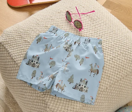 Kinder-Boardshorts mit Rittermuster liegen auf einem Hocker, daneben ein Skateboard, eine Sonnenbrille und ein Ball.