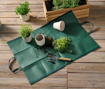 Eine Gartenabfalltasche liegt auf einem Holzboden neben Topfpflanzen und einer Gießkanne.